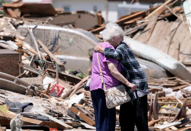Two older adults hug in front of a destroyed home. One holds a cane.