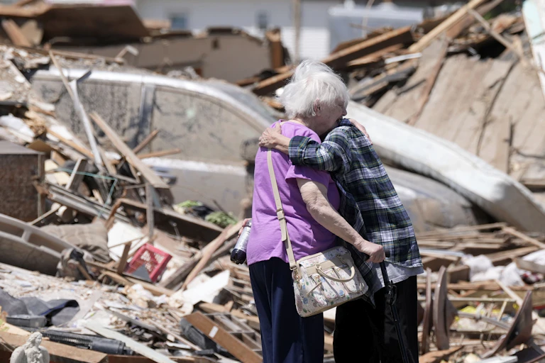 Two older adults hug in front of a destroyed home. One holds a cane.