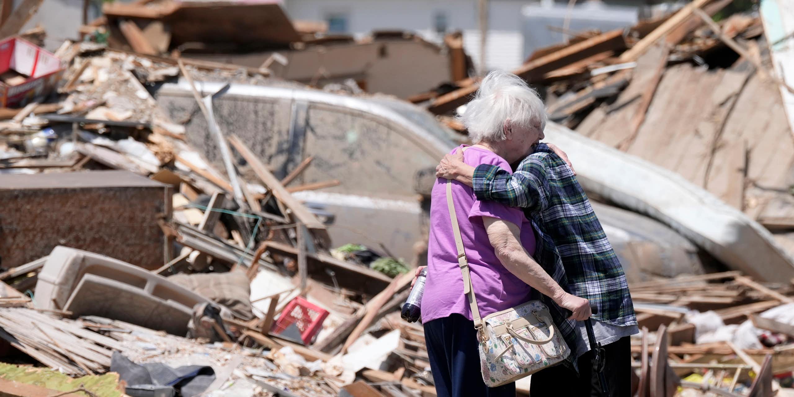 Two older adults hug in front of a destroyed home. One holds a cane.