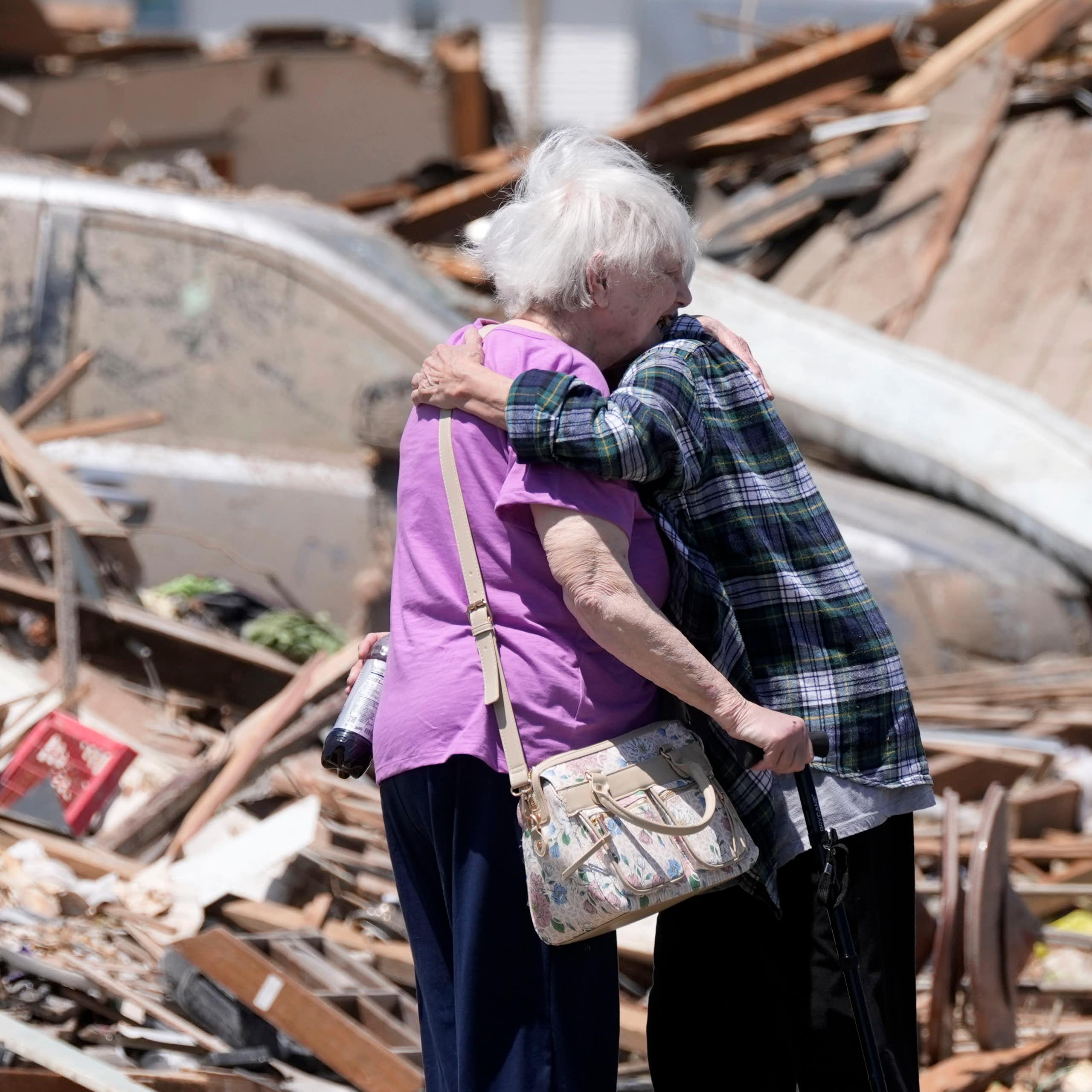 Two older adults hug in front of a destroyed home. One holds a cane.