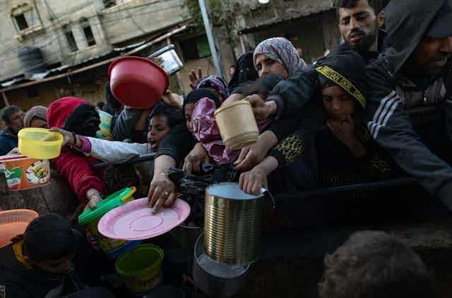 A crowd of Palestinian people holding plates and containers as they wait to collect food.