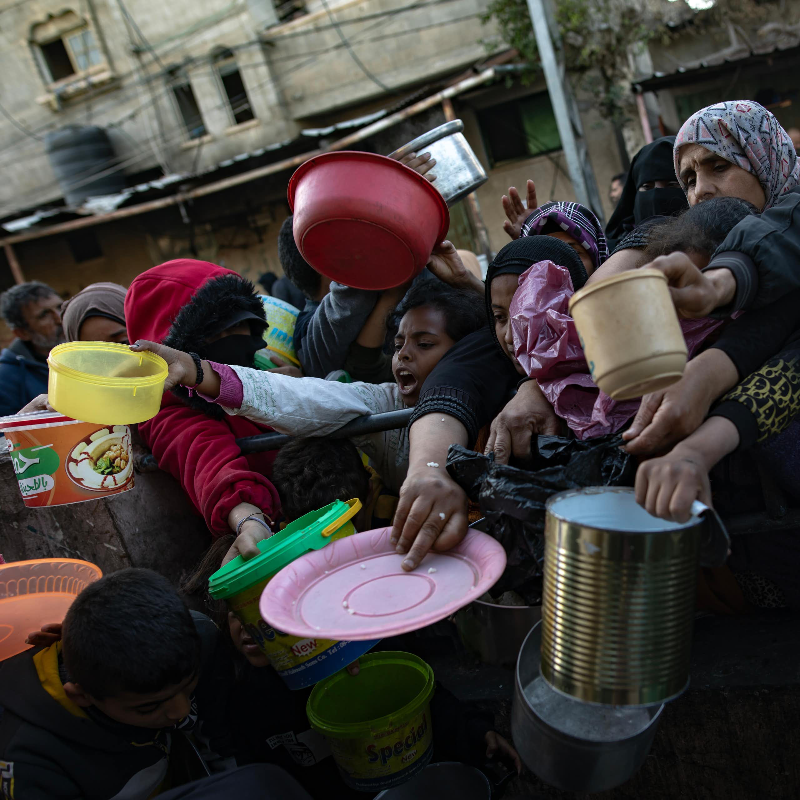 A crowd of Palestinian people holding plates and containers as they wait to collect food.