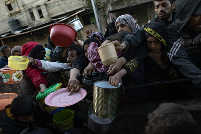 A crowd of Palestinian people holding plates and containers as they wait to collect food.