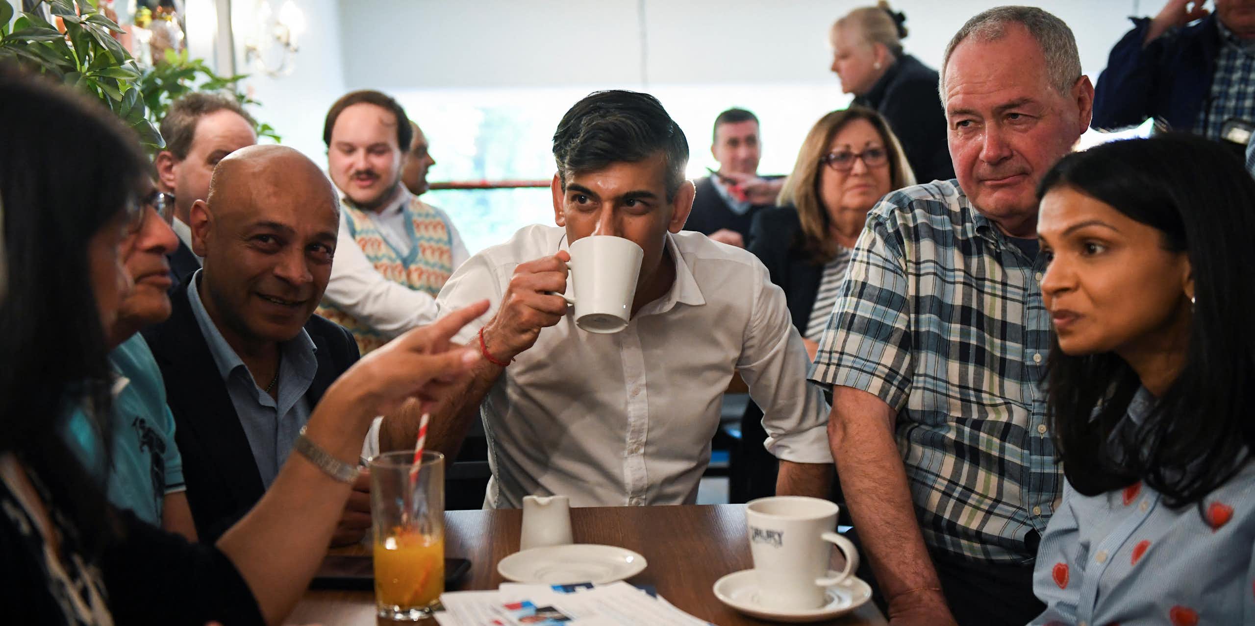 A man in a white shirt drinks a cup of tea surrounded by other people.