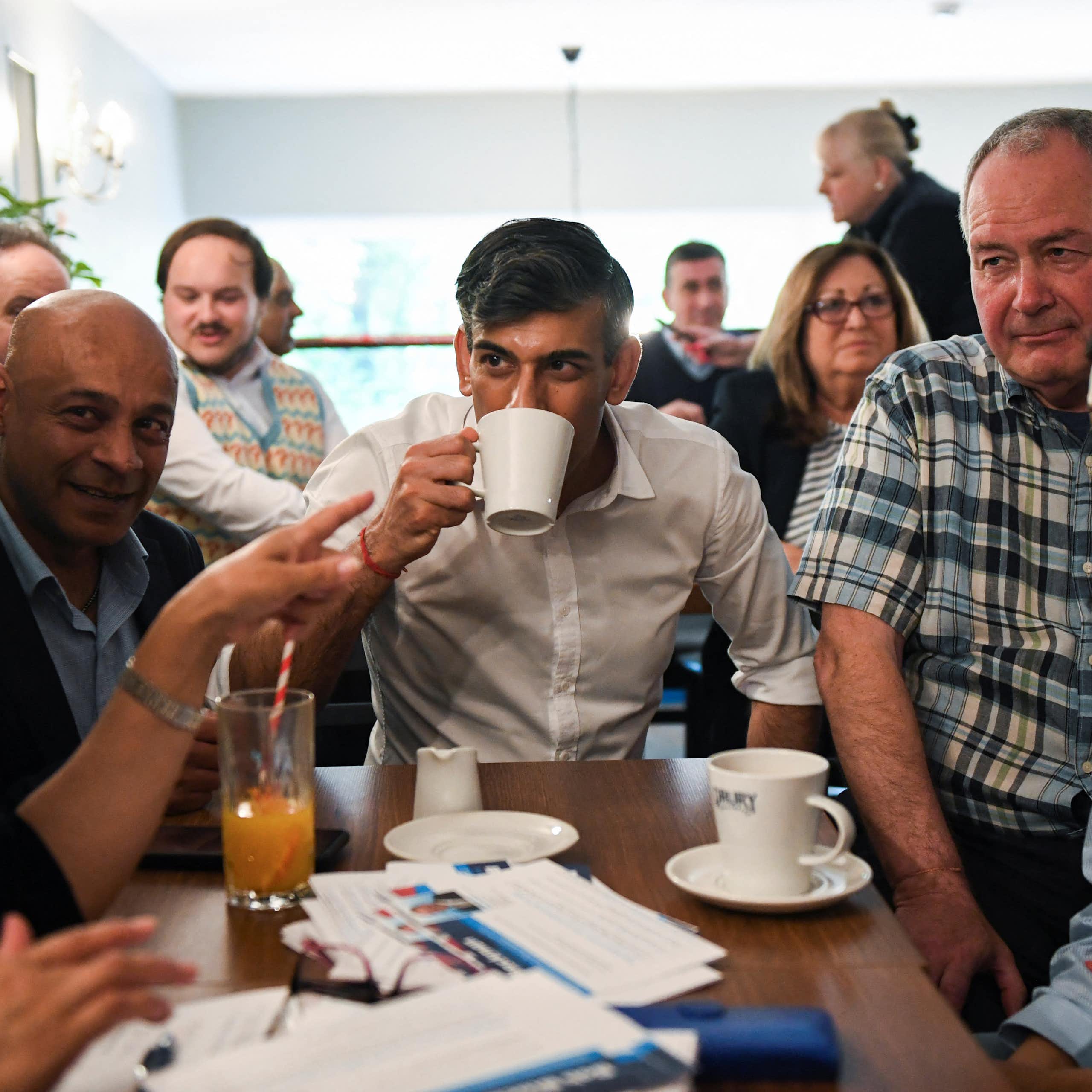 A man in a white shirt drinks a cup of tea surrounded by other people.