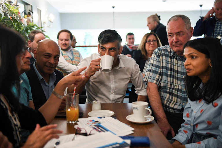 A man in a white shirt drinks a cup of tea surrounded by other people.