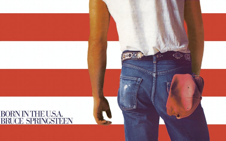 Man wearing sleeveless white t-shirt tucked into blue jeans, viewed from behind.