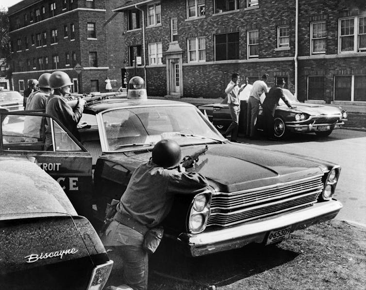 Several policemen point their weapons at Black suspects who have their hands on a parked car.