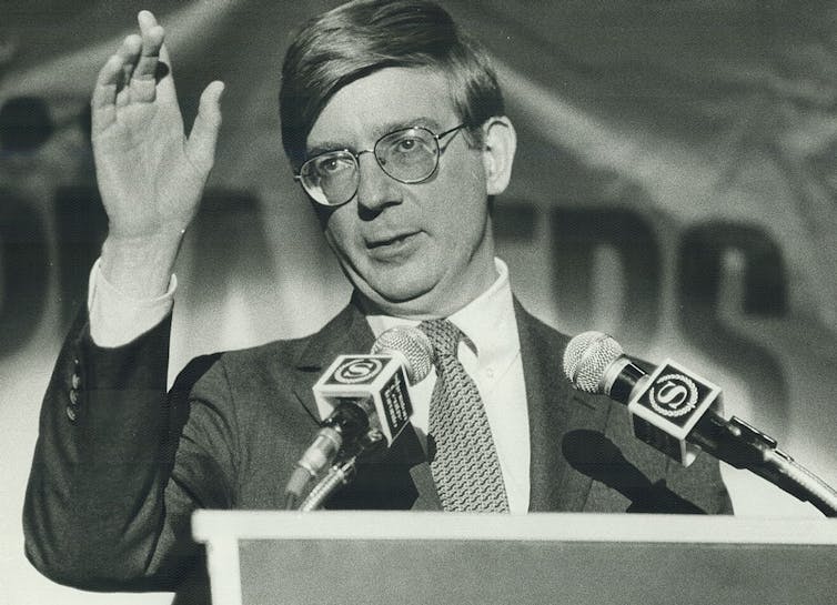Black and white photograph of a man with a toupee haircut and glasses gesturing while speaking at a lectern.