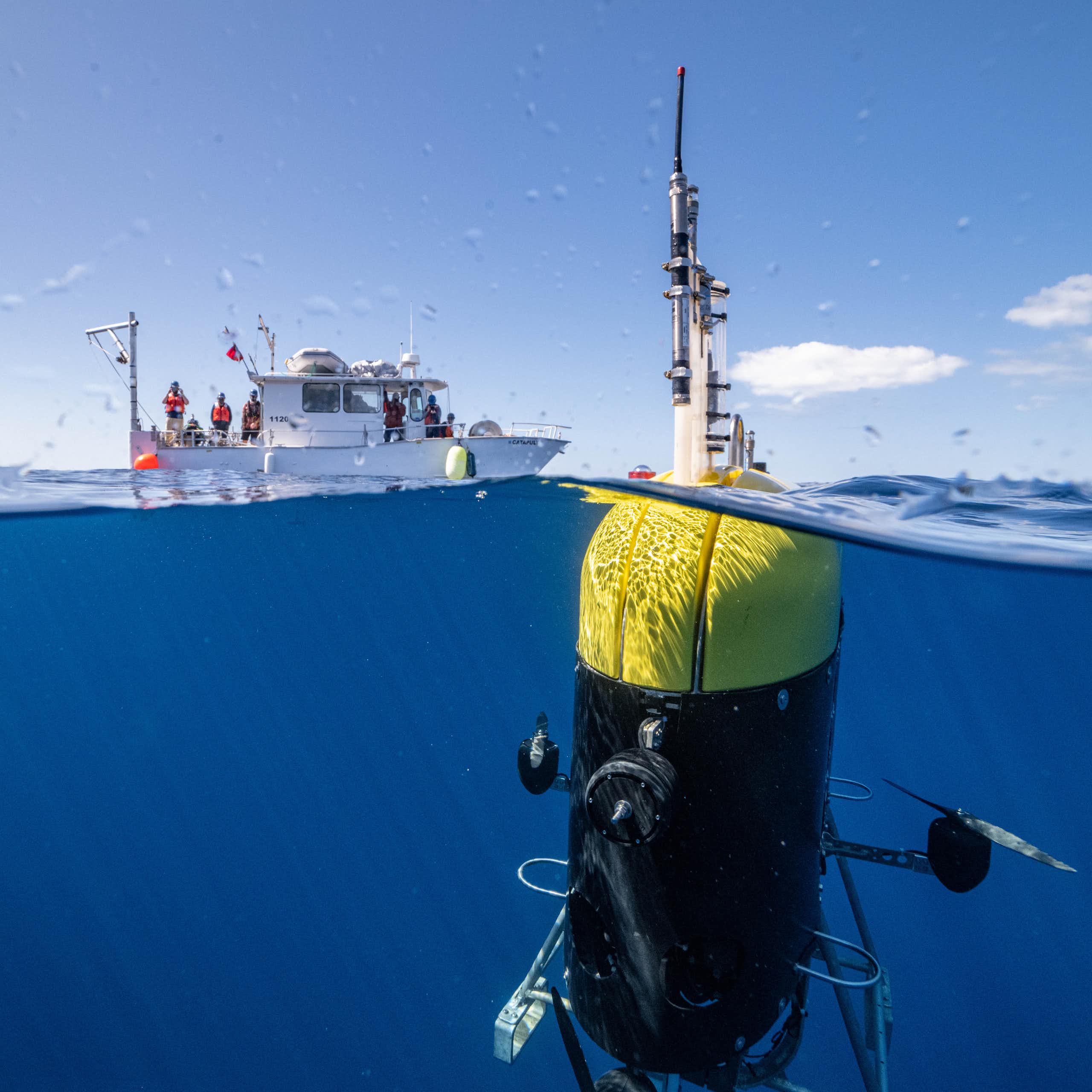 Un gran robot cargado de sensores aparece con su antena por encima del agua y el resto bajo la superficie, con un barco de investigación al fondo.