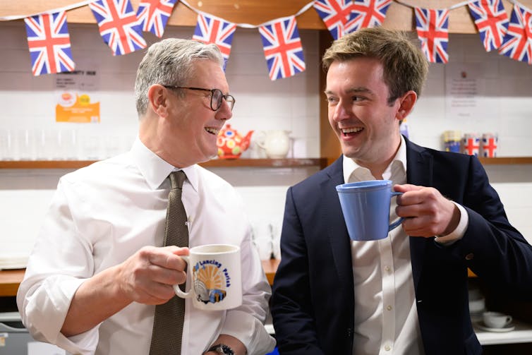A man in a white shirt chats to a man in a blazer. Bith hold cups.