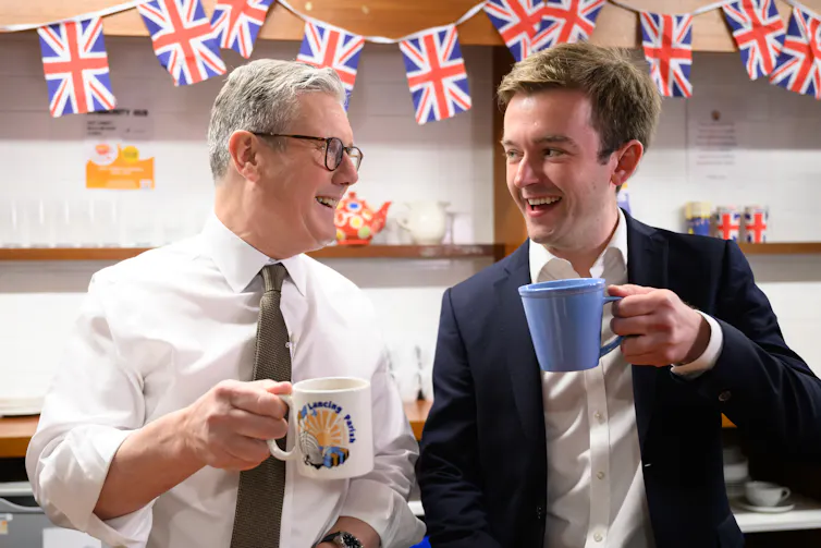 A man in a white shirt chats to a man in a blazer. Bith hold cups.