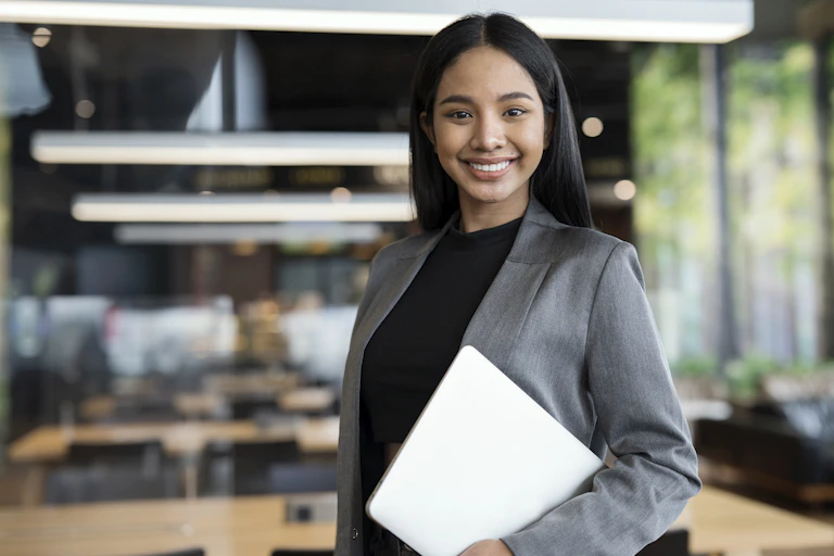 A young woman holding a laptop and wearing a gray blazer smiles as a series of table work stations stand in the background.