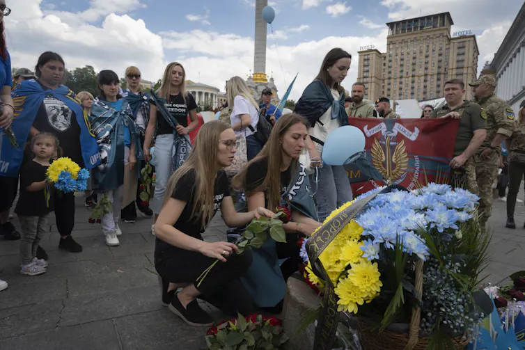 Des femmes déposent des fleurs au milieu d’une rue, entourée par une foule