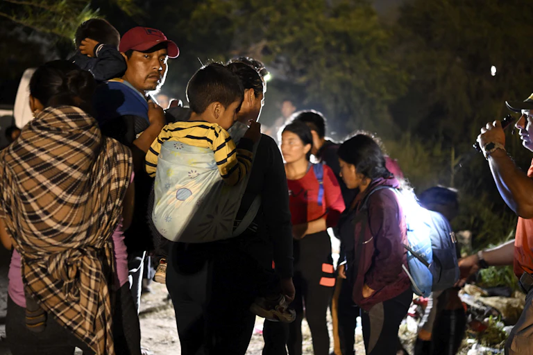 A man wearing a baseball cap talks with a crowd of women and children.