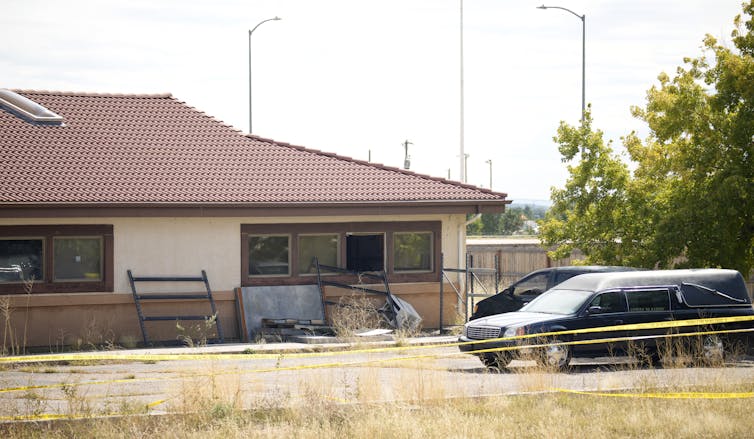 Two cars sit behind crime scene tape in front of a single story building with a tile roof