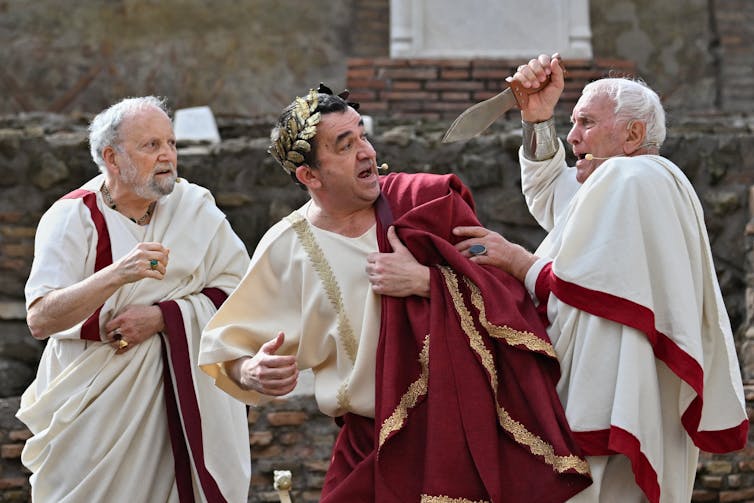 Men in purple-trimmed togas wrestle with each other, one swings a knife, another stares in horror.
