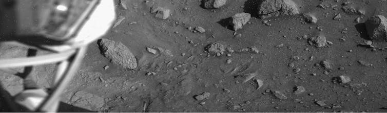 Black and white photo of a rocky floor with struts from the lander's surface sample arm housing