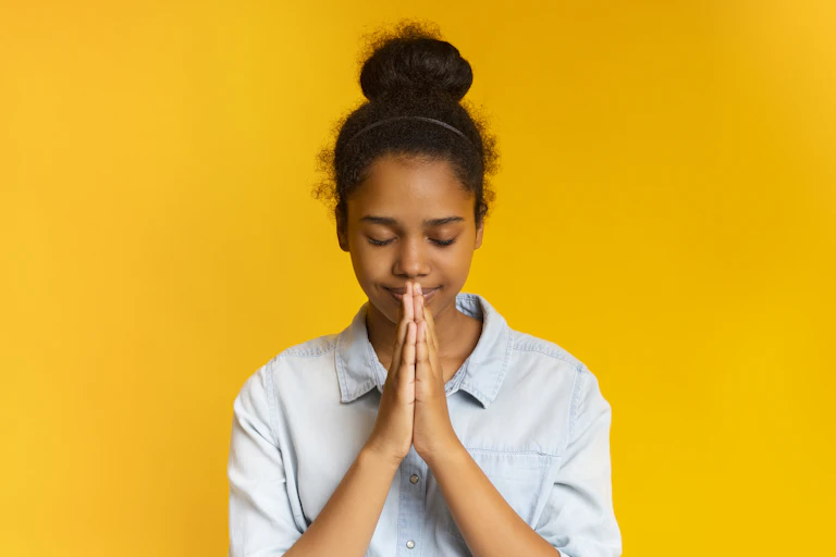 An adolescent girl with her hair in a bun prays with her hands clasped. The background is yellow.