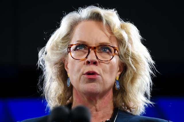 A blonde woman with glasses speaks at a podium