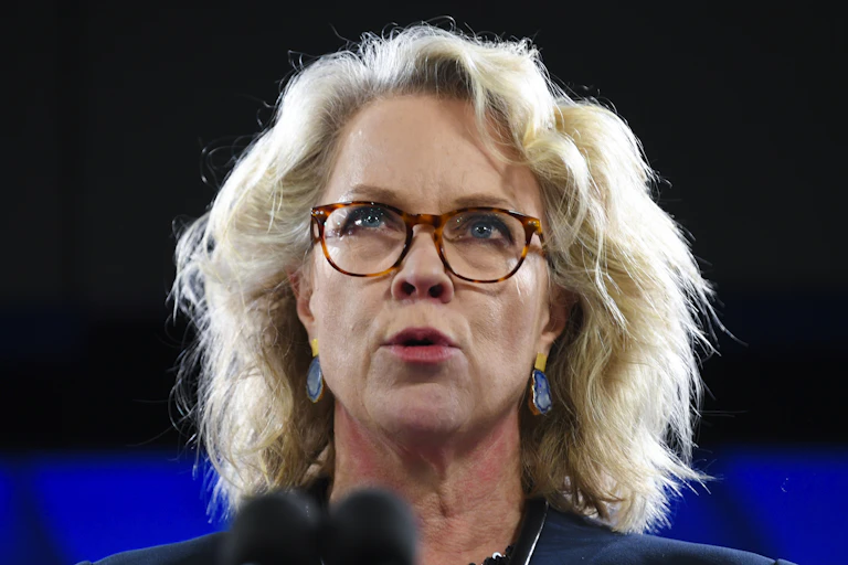A blonde woman with glasses speaks at a podium