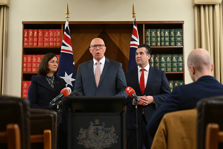 A bald man with glasses stands behind a lectern.A woman stands to his left and a man on his right, with two Australian flags behind them all