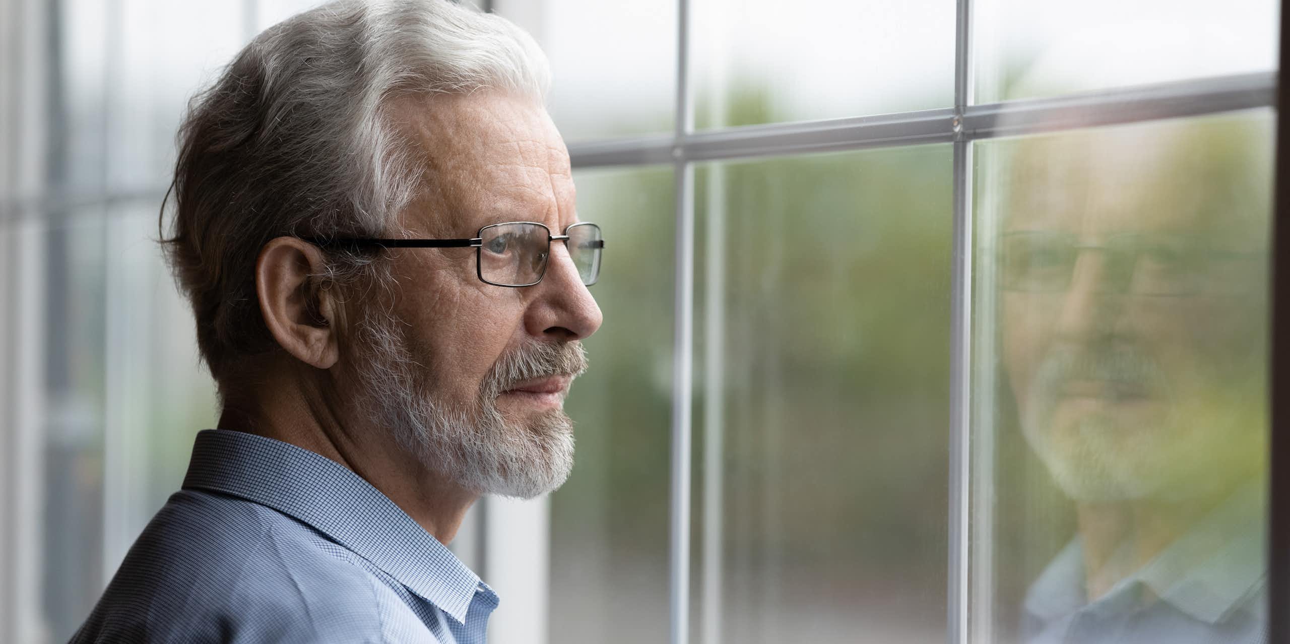 older man is looking out window and his reflection is seen in the glass