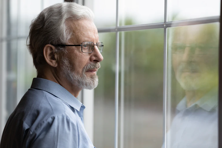 older man is looking out window and his reflection is seen in the glass