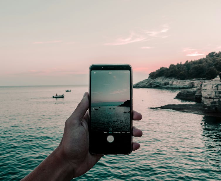 a hand holding a phone up to take a photo of a rocky bay