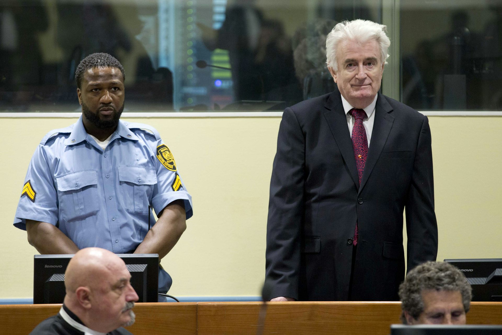 A man with white hair dressed in a suit standing in a courtroom next to a guard.