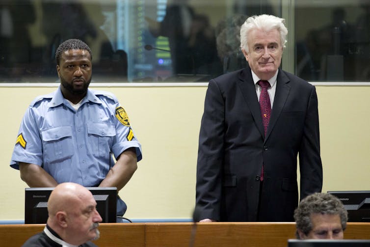A man with white hair dressed in a suit standing in a courtroom next to a guard.