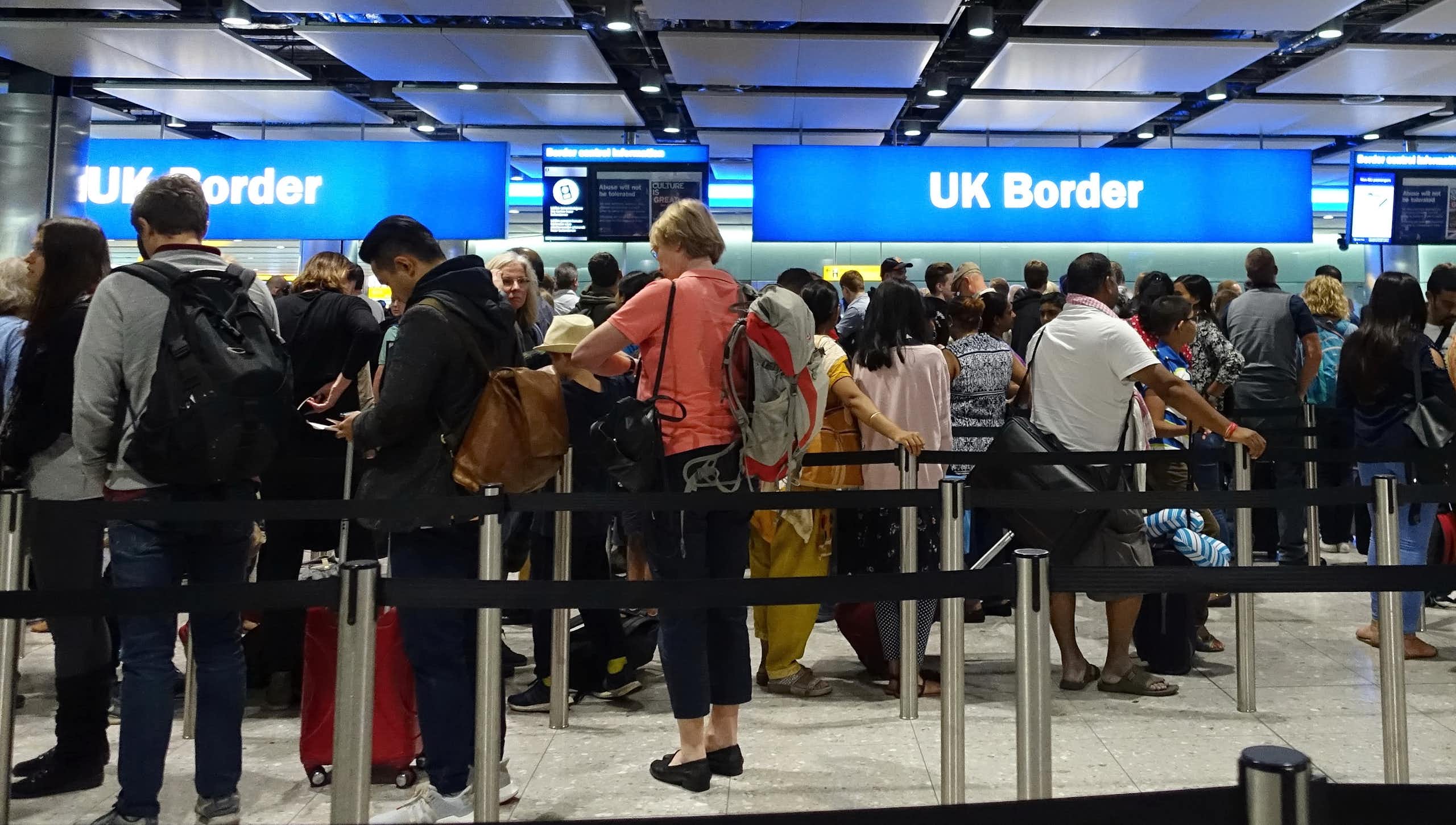 Photo of queues of travelers lining up at the glowing blue UK border sign in an airport