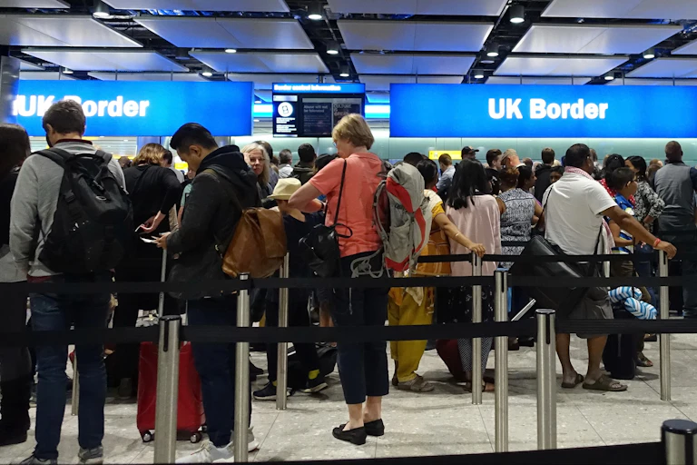 Photo of queues of travelers lining up at the glowing blue UK border sign in an airport