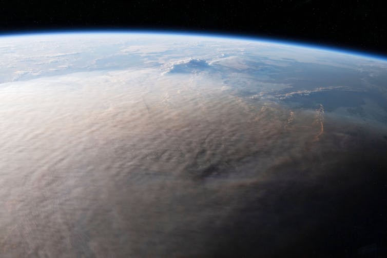 A high altitude view of Earth with its curve clearly visible and a brown grey plume covering most of the visible surface.