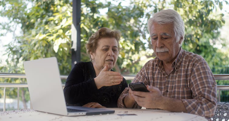An elderly woman and an elderly man are sitting outside at a table with an open laptop in front of them.