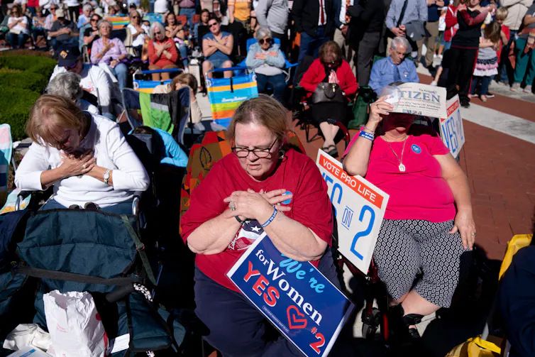 People sit in folding chairs in a row and cross their arms and look down. One holds a poster that says 'We're women for yes.'