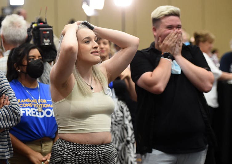 People stand together and look surprised, as one covers their mouths and one has her hands in her hair.