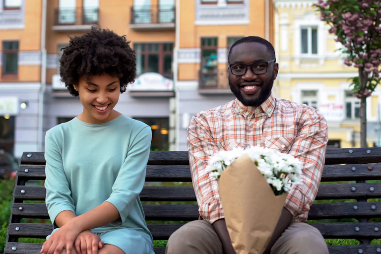 Un homme et une femme sont assis sur un banc public et sourient. L'homme porte un bouquet de fleurs