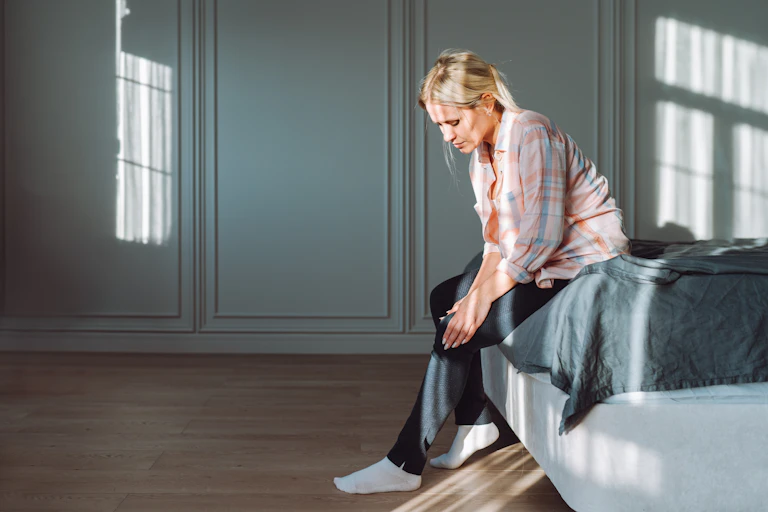 A middle-aged woman sits on the edge of her bed, hunched over and holding her knee as if in pain.