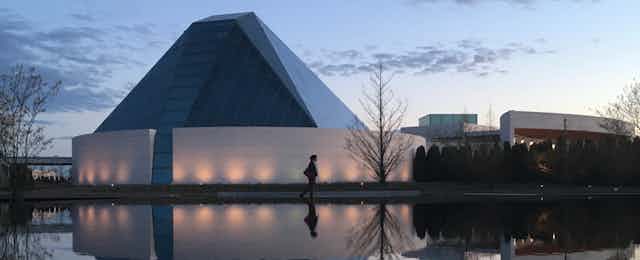 A woman walks along a path near a body of water. behind her is a conical dome structure. 