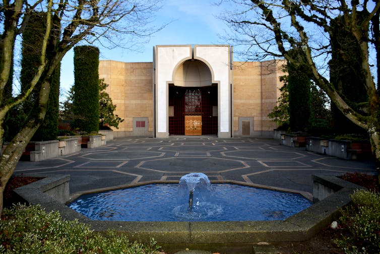 A mosque courtyard with trees and a fountain. The courtyard stone features a geometric pattern.