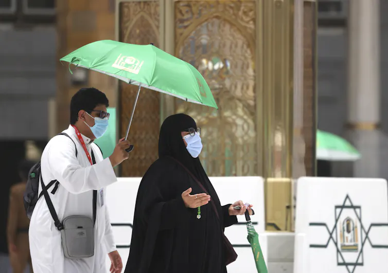 A man dressed in a white robe stands behind a women dressed in black as she raises her hands reciting a prayer.