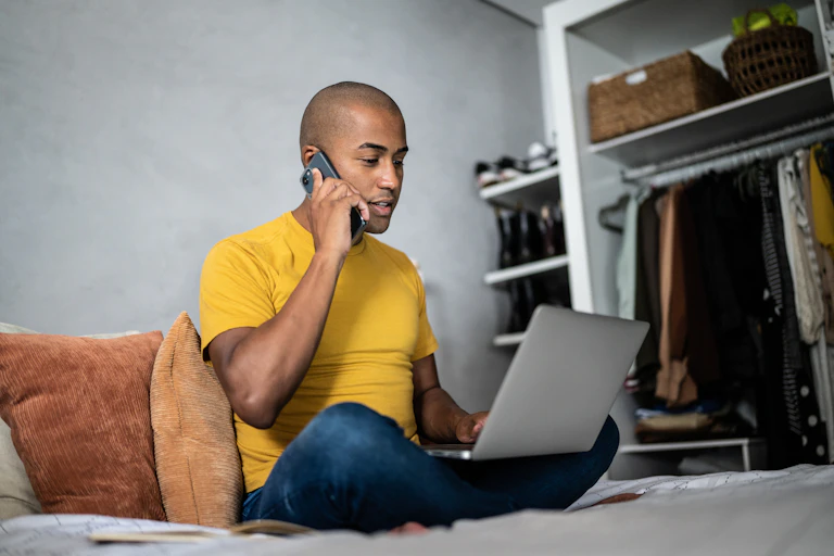 A young man with an engaged expression speaks on a cellphone with a laptop open in front of him.