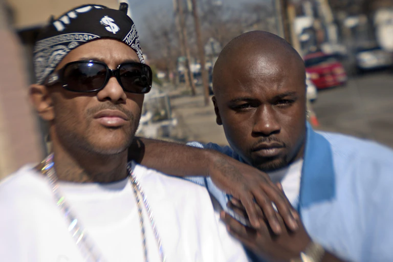 Two Black men pose for a photo on a sidewalk.