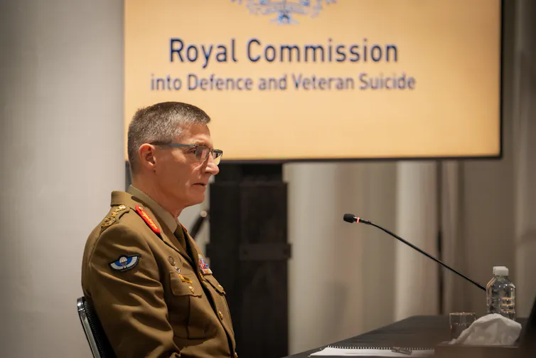 A man in an arm uniform sits in front of a screen saying Royal Commission into Defence and Veteran Suicides