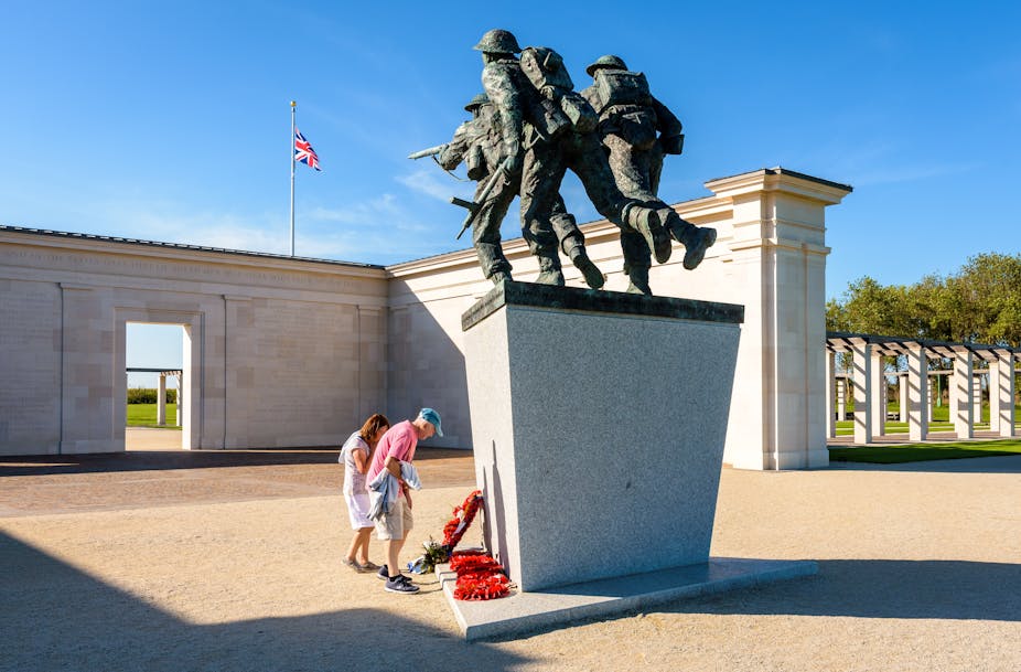 Deux personnes se recueillent au pied d'un monument commémoratif. Derrière, on voit le drapeau britannique.