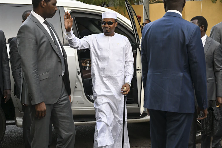 Man wearing white traditional dress alights from a vehicle surrounded by men wearing suits