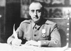 A black-and-white photo shows a man in an uniform holding a quill as he signs a paper on the desk he's sitting at.