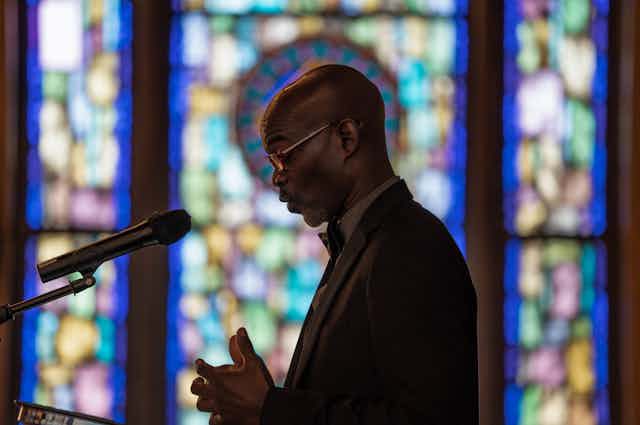 A Black man in glasses and a suit looks down as he speaks into a microphone, standing in front of stained glass windows.