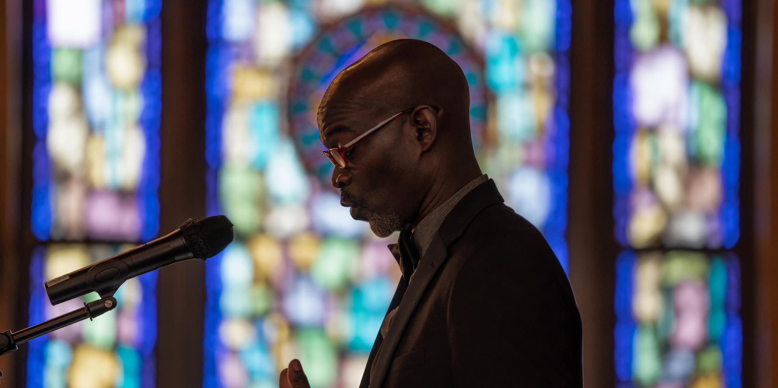 A Black man in glasses and a suit looks down as he speaks into a microphone, standing in front of stained glass windows.